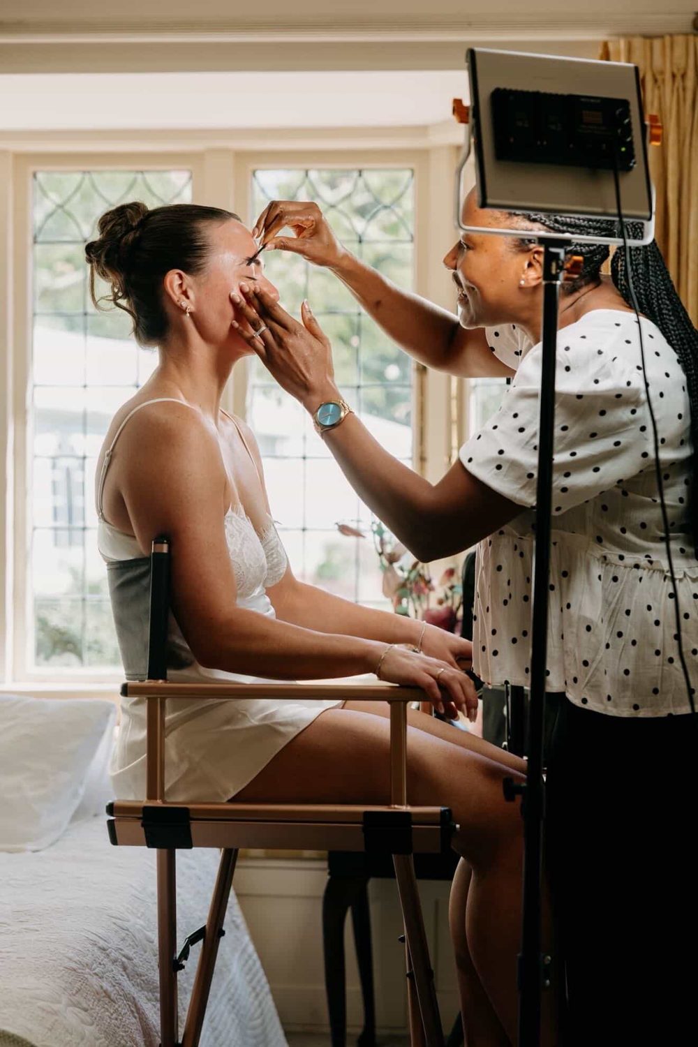 Emande applying a bride's makeup by the window on her wedding morning